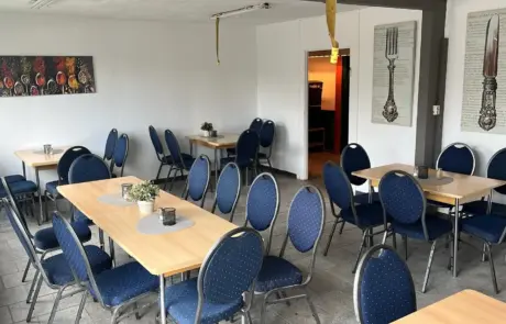 Bright cafeteria with blue chairs and wooden tables in Gewerbegebiet Altlandsberg, featuring framed cutlery art and a service doorway.
