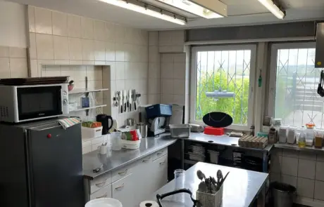 In der Kantine im Gewerbegebiet Altlandsberg stehen Edelstahlarbeitsflächen, Küchenutensilien, ein Kühlschrank und mehrere Geräte neben den Fenstern.